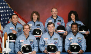 The seven members of the Challenger Crew pose, smiling, in front of the American flag with their space helmets on their laps.