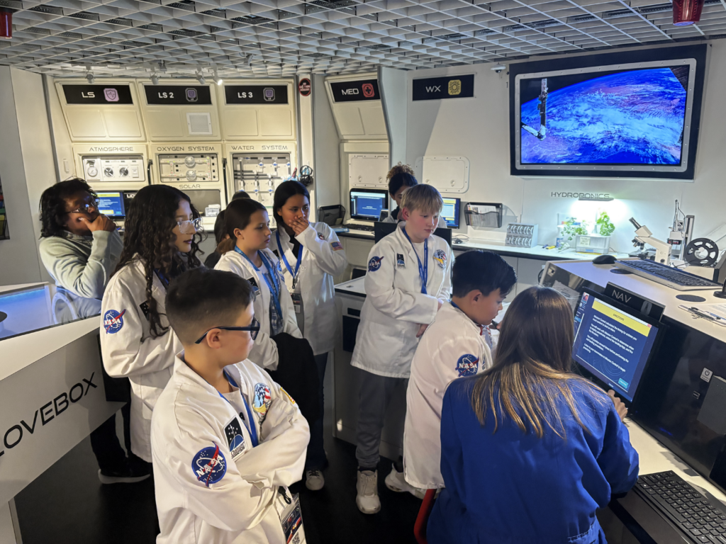 Students in lab coats stand in a simulated spacecraft, looking at a computer screen.